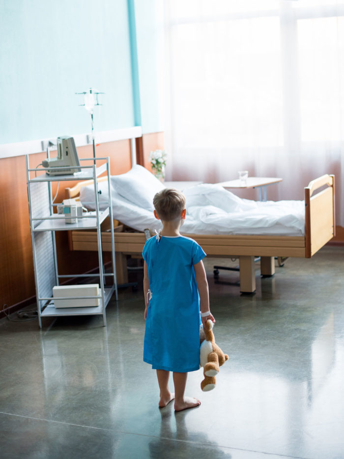Back view of little barefoot boy with teddy bear standing in hospital room