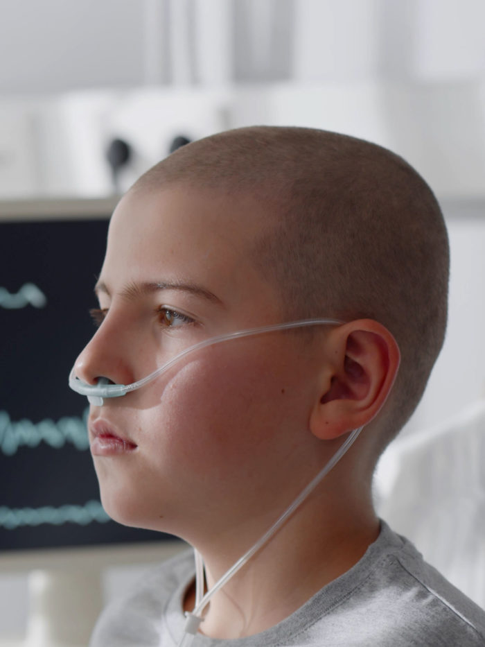 Side view of teen boy with cancer sitting in bed with vital signs on computer monitor