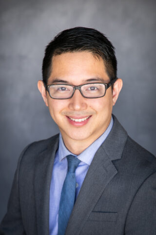 Headshot Dr. Alexander Tan - Man wearing suit and glasses smiles into camera in front of blue background