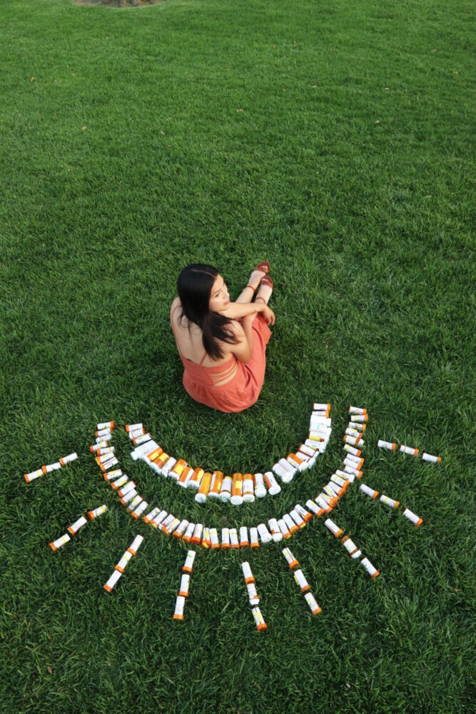 Liliana sitting in the grass with prescription medications arranged in a sun around her